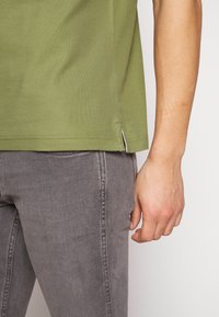 Chemise verte à manches courtes avec un ourlet droit et des fentes latérales, associée à un pantalon en denim gris avec des coutures visibles et une coupe slim.