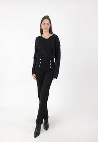Donna in piedi che indossa un maglione nero a V, pantaloni neri a vita alta con bottoni e stivaletti neri a punta su sfondo bianco.