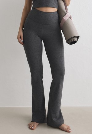 Kvinde iført mørkegrå tætsiddende yogabukser og en matchende cropped top, der holder en sammenrullet lyserød yogamåtte med skulderrem.