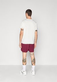 Camisa blanca de manga corta con ribetes en contraste, combinada con bermudas burdeos. El modelo lleva zapatillas blancas y calcetines tobilleros; tatuajes visibles en las piernas.