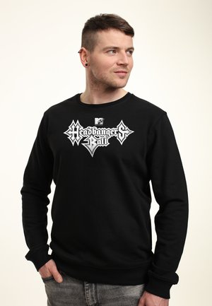 Uomo con capelli corti che indossa una felpa nera con il logo bianco "MTV Headbangers Ball", guarda verso destra, mani nelle tasche dei jeans grigi.