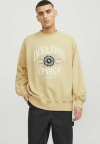 Sweatshirt beige à manches longues, avec des bords côtelés aux poignets et au col. Présente un texte imprimé blanc et un design de logo sur le devant. Associé à un pantalon sombre.