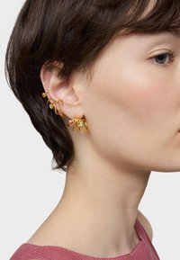 Boucles d'oreilles multiples en ton or, avec divers designs, mettant en avant des pierres précieuses colorées. Le métal texturé et les formes variées ornent l'oreille.