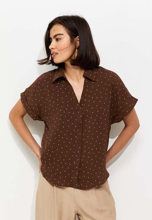 Kobieta o ciemnych włosach do ramion, ubrana w brązową, krótką koszulę na guziki w białe grochy i beżowe spodnie, patrząca w lewo.