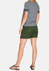Camisa gris de manga corta con mangas texturizadas, combinada con pantalones cortos verdes. El atuendo presenta un ajuste casual y sandalias planas.