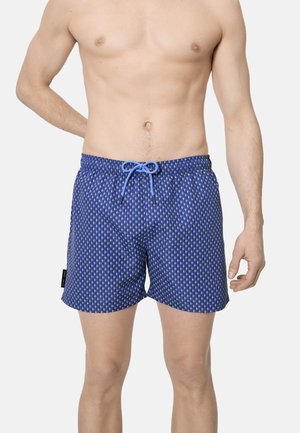 Modelo masculino vistiendo bañador azul con estampado y cintura con cordón, posando frente a un fondo claro y liso.