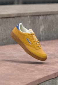 Žute i plave Reebok tenisice lebde iznad betonskih stepenica, prikazujući bočni logo i gumenog potplat u vanjskom okruženju.