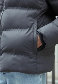 Purus pilkas švarkas su horizontaliai siuvinėtomis dalimis, su šonine kišene ir elastingomis rankovėmis, pagamintas iš lygios, vandeniui atsparios medžiagos.