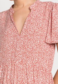Rotes Blumenkleid mit V-Ausschnitt, kurzen Puffärmeln und elastischem Taillenbund. Der Stoff zeigt ein weißes Blättermuster auf einem strukturierten Hintergrund.