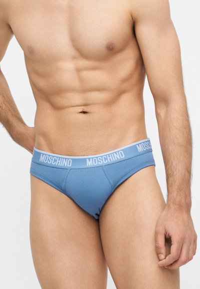 Ανδρικός κορμός που φοράει ανοιχτό μπλε εσώρουχα Moschino τύπου σλιπ με λογότυπο στη μέση, στέκεται μπροστά σε ομοιόμορφο φόντο.