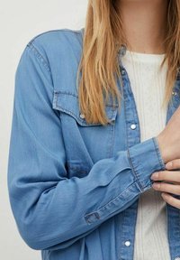 Chemise en denim bleu clair à manches longues, poignets boutonnés, et deux poches poitrine. Caractérisée par une texture douce et des détails de couture subtils.