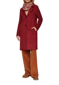 Manteau en laine rouge au design ajusté, fermeture à un bouton, et poches latérales, associé à une écharpe à carreaux et à un pantalon en velours côtelé marron.