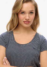 Ung kvinde med blondt hår iført en navyblå og hvidstribet kortærmet skjorte, med armene krydset, kigger en smule væk fra kameraet.