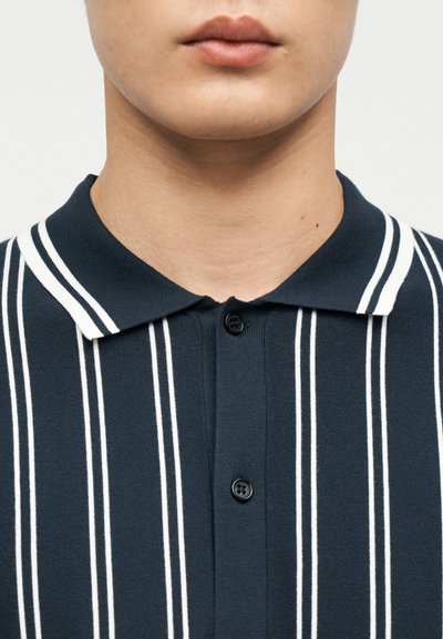 Εγγύτερη λήψη του κατώτερου μέρους προσώπου και λαιμού ενός ατόμου που φοράει σκούρο μπλε πόλο πουκάμισο με λευκές κάθετες ρίγες και δύο μαύρα κουμπιά κουμπωμένα.