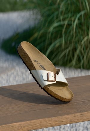 Hvid Birkenstock-sandal med enkelt rem, korksål og metalspænde, der hviler på en træoverflade udendørs med grønt område i baggrunden.