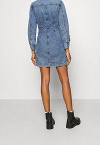 Jeansové šaty s dlouhými rukávy, přiléhavý design a zadními záševky. Nosené s černými kotníkovými botami. Hladká textura a středně modrá barva.