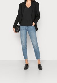 Blazer nero sopra una maglietta nera a V, abbinato a jeans tapered blu chiaro e mules nere con tacco. Borsa strutturata tenuta in una mano.