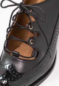 Chaussure brogue en cuir noir avec détails brogue, intérieur beige, et lacets noirs uniques bouclés passant à travers des œillets dorés.
