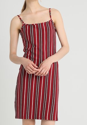 Vrouw die een mouwloze, knielange rode jurk met verticale zwarte en witte strepen draagt, staand met de handen gevouwen voor zich.