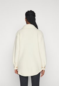 Crème, textuur fleece jas met een kraag, een ronde zoom en ronde knopen op de manchetten. Achteraan zichtbaar met een ontspannen pasvorm.