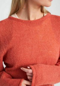 Adīta džemperis siltā korallkrāsā. Iezīmējas ar garām piedurknēm ar izstieptiem reģipšiem un platu kakla izgriezumu. Mīksta, strukturēta vilnas auduma.