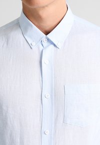 Ljusblå linneskjorta med krage med knappar, fyra knappar fram och en enskild bröstficka, med en strukturerad tyg.