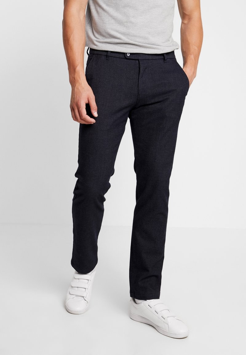 Pantaloni navy in tessuto texturizzato e slim fit, con chiusura a pulsante e tasche laterali, abbinati a sneaker bianche.