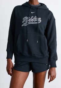 Svart Nike-hoodie med framficka, dragsko och logotyp i vitt med en strukturerad finish. Matchas med svarta shorts, båda i ett mjukt tyg.