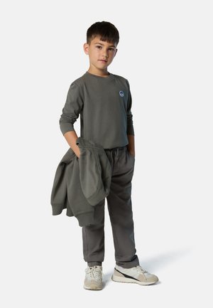 Graues Langarmshirt mit rundem Logo, passende graue Hose und hellfarbige Sneaker. Der Junge steht mit den Händen in den Taschen und hält einen Sweatshirt.