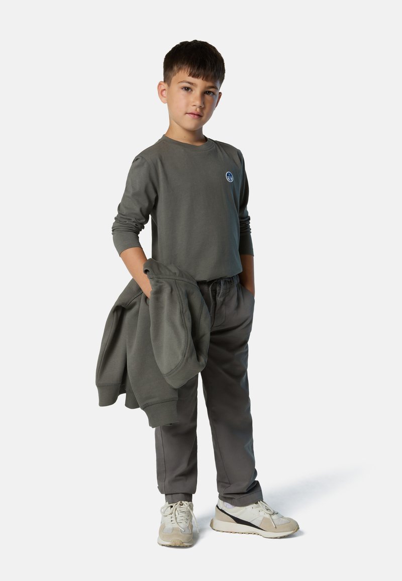 Camisa gris de manga larga con un logo redondo, pantalones grises a juego y zapatillas de color claro. El chico está de pie con las manos en los bolsillos y sostiene una sudadera.