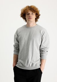 Sudadera gris con cuello redondo acanalado, mangas largas y puños y dobladillo acanalados. Textura suave y corte relajado, combinada con pantalones negros.