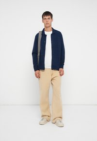 Cárdigan de malha azul-marinho com detalhe cinza, frente com botões e bainha canelada. Combinado com uma camisa em tom off-white e calças largas em bege claro.