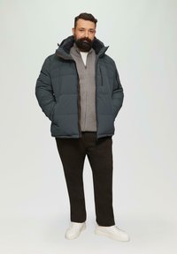 Grijze gewatteerde pufferjack met een capuchon, rits aan de voorkant en zijzakken, gedragen over een grijze trui en donkere broek, compleet met witte sneakers.