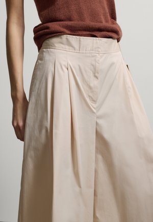 Primer plano de una persona que lleva puestos pantalones anchos beige de cintura alta con pliegues delanteros y una camiseta sin mangas marrón metida por dentro.