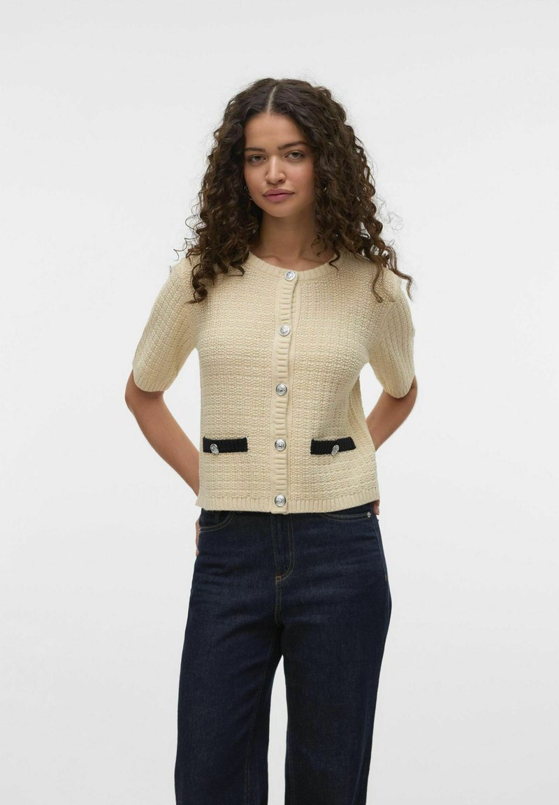 Cardigan en tricot beige à manches courtes, avec un motif texturé, de grands boutons argentés et des accents de poche noirs.