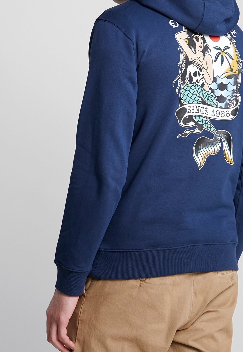 Personne portant un sweat à capuche bleu marine avec un motif coloré représentant une sirène, un crâne, un palmier et des vagues, avec le texte « Since 1966 » dans le dos.