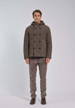 Hombre de pie con una chaqueta marrón de doble botonadura y textura con capucha, pantalones de pana grises y zapatos de cuero marrón sobre un fondo sencillo.