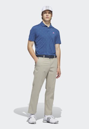 Male model wearing white cap, blue patterned polo shirt, beige pants, navy belt, and white golf shoes standing against a plain background.