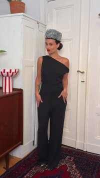 Γυναίκα με μαύρη ολόσωμη φόρμα με έναν ώμο, φοράει ασημένιο γλυπτό κεφαλόδεσμο και στέκεται μέσα σε κλειστό χώρο δίπλα σε ντουλάπι και κόκκινο μοτίβο χαλί.