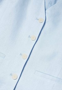 Chemise en lin bleu clair avec un col à revers, deux poches avant et quatre boutons ronds couleur crème avec des coutures visibles.