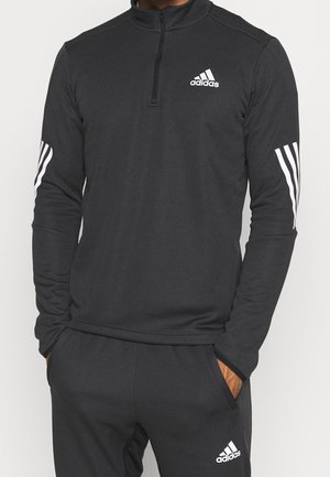Czarny dres Adidas z białym logo na klatce piersiowej i udzie, białe paski na rękawach, noszony przez osobę z rękami w kieszeniach.