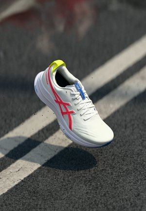 Weißer Sportschuh mit rotem Logo, neon-gelber Ferseneinsatz und blauen Details, schwebt über strukturiertem Asphalt mit weißen Fahrbahnmarkierungen.