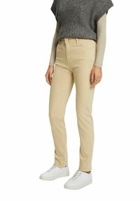 Jeans de tiro alto en beige con un corte ajustado y un diseño clásico de cinco bolsillos. Combinados con una blusa de punto gris texturizada y zapatillas blancas.