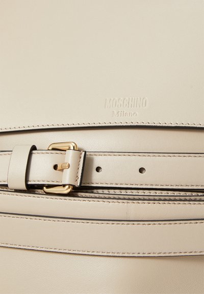 Beige ādas soma ar zelta toni sprādzi, ar izspiedu "MOSCHINO Milano" logotipu un kontrastējošām šuvēm pa malām.