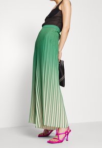 Frau, die einen gefalteten Rock in Grün- und Creme-Ombre trägt, ein schwarzes ärmelloses Oberteil, leuchtend pinke Riemchensandalen und eine schwarze Clutch-Tasche hält.