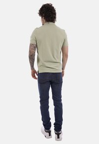 Hombre con cabello rizado y brazo derecho tatuado, vistiendo una camiseta tipo polo verde claro, jeans azul oscuro y zapatillas blancas, de espaldas sobre un fondo blanco.