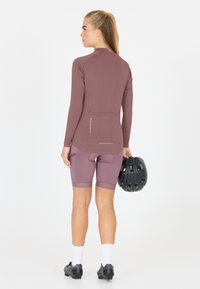 Vrouw in mauve fietsoutfit die een zwarte helm vasthoudt, van achteren gezien, staand tegen een witte achtergrond.