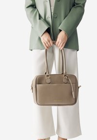 Una persona indossa un blazer verde chiaro e pantaloni bianchi, mentre tiene una borsa in pelle taupe con manici doppi e dettagli in oro.