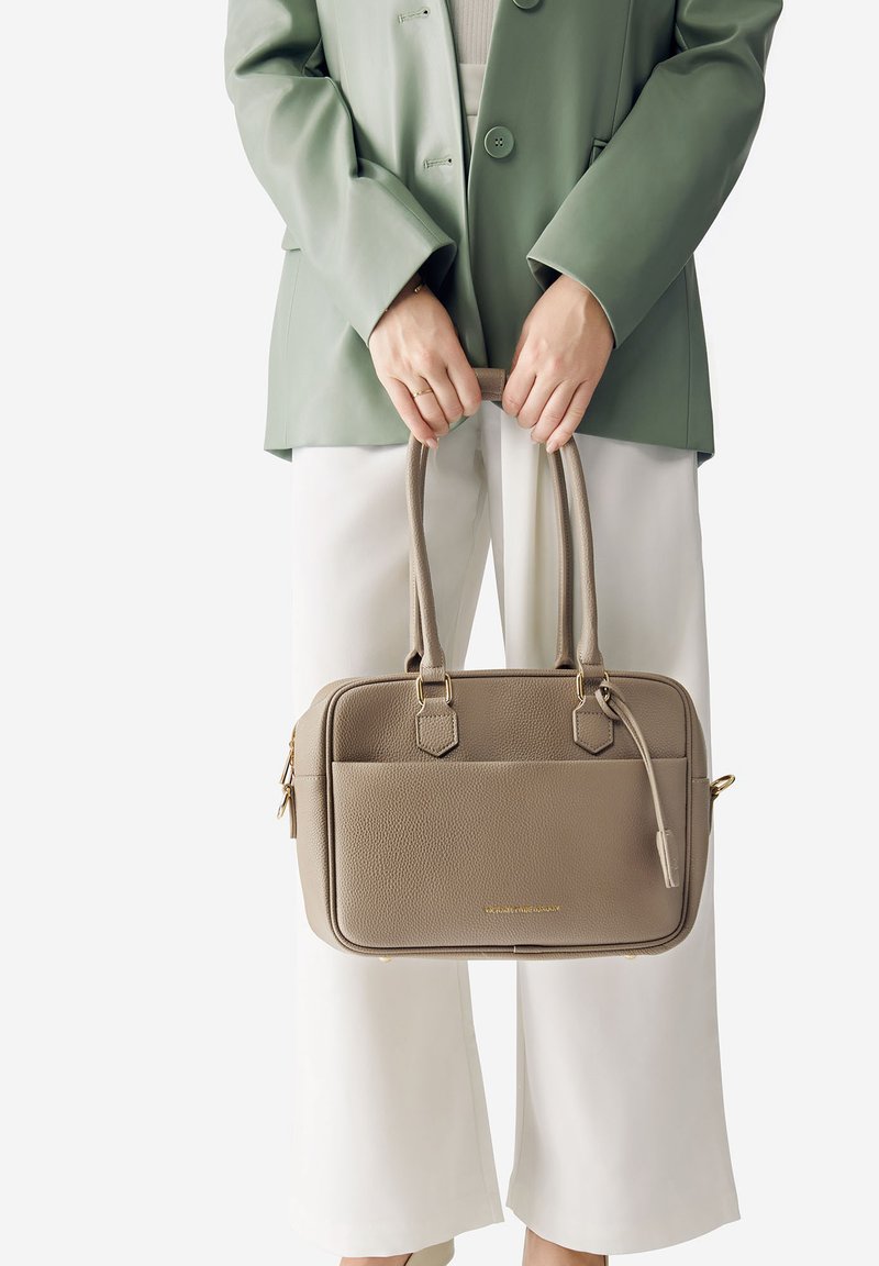 Una persona indossa un blazer verde chiaro e pantaloni bianchi, mentre tiene una borsa in pelle taupe con manici doppi e dettagli in oro.