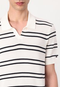 Hvit strikket polo skjorte med svarte horisontale striper. Har V-hals og korte ermer, med et logoakcent på ermet.